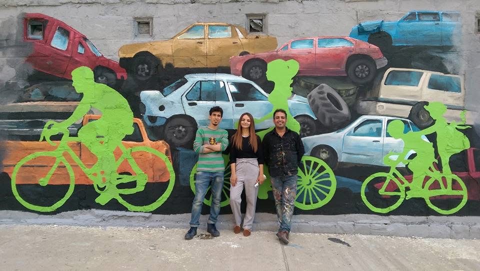 Outdoor wall mural with stacked painted cars and bright green cyclist silhouettes, with three people standing in front.